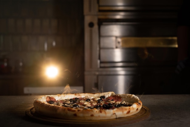 Salami pizza in an Italian restaurant on a wooden table. In the background is a backlit oven. Hot baked goods.