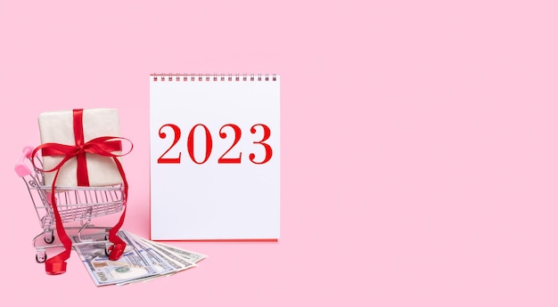 Sale of 2023 Cart with gift box with red bow and cash on pink background with calendar date 2023 Buying gifts