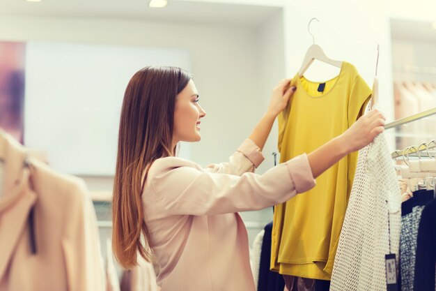 продажа, одежда, шопинг, мода и люди концепция - счастливая молодая женщина выбирает между двумя рубашками в торговом центре или магазине одежды