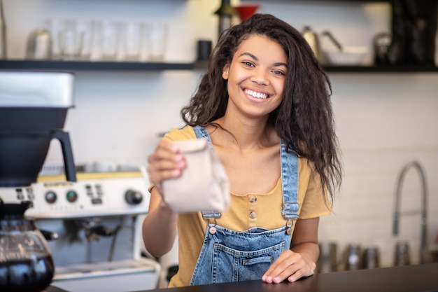 Sale, coffee. Cheerful smiling young woman in denim overalls standing behind counter of coffee shop holding out paper bag