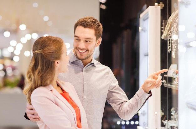 sale, consumerism and people concept - happy couple pointing finger to shopping window at jewelry store in mall