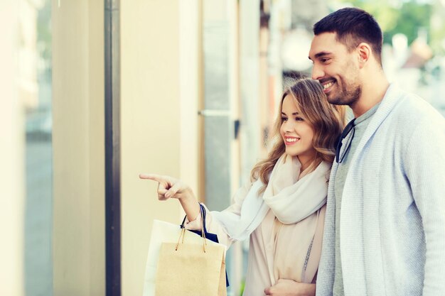 Photo sale, consumerism and people concept - happy couple with shopping bags pointing finger to shop window on city street