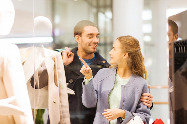 Vendita, consumismo e concetto di persone - giovane coppia felice con borse della spesa che punta il dito alla vetrina del negozio nel centro commerciale