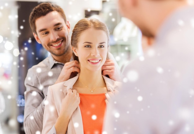 sale, consumerism, presents, holidays and people concept - happy couple trying golden pendant on at jewelry store in mall with snow effect