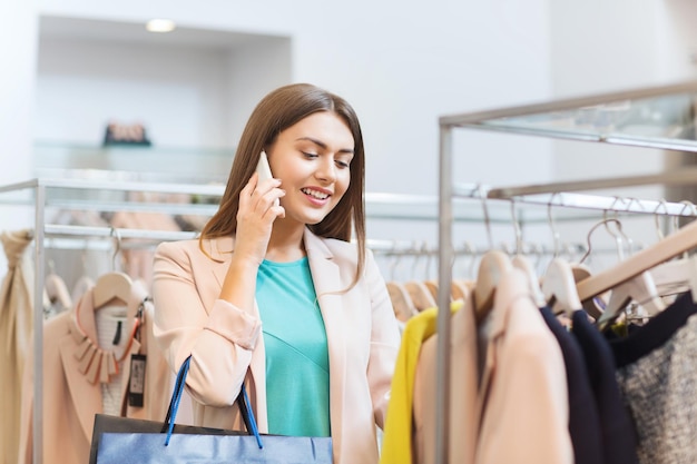 sale, consumerism, shopping and people concept - happy young woman with shopping bags choosing clothes and calling on smartphone in mall or clothing store