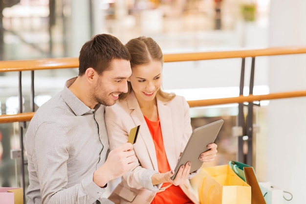 sale, consumerism, technology and people concept - happy young couple with shopping bags and credit card pointing finger to tablet pc computer in mall