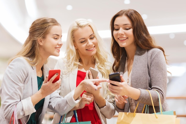 sale, consumerism, technology and people concept - happy young women with smartphones and shopping bags in mall