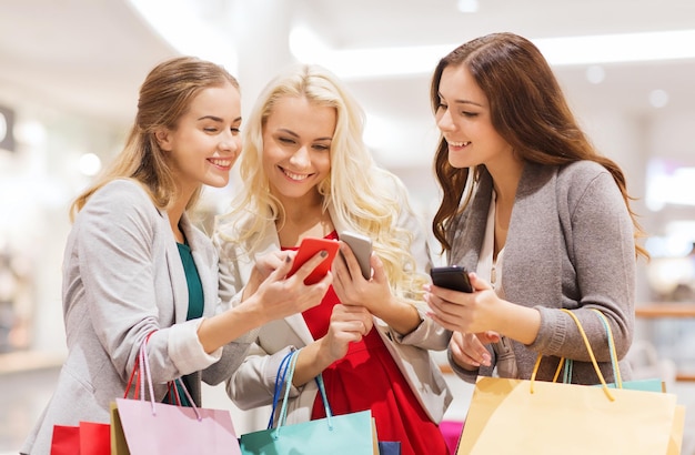 sale, consumerism, technology and people concept - happy young women with smartphones and shopping bags in mall