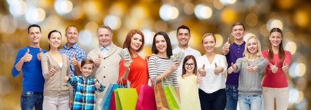 sale, family, generation and people concept - group of happy men and women with shopping bags and money showing thumbs up over holidays lights background