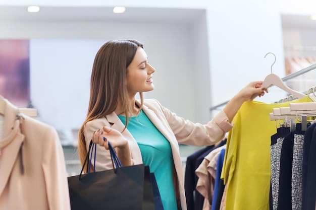 sale, fashion, consumerism and people concept - happy young woman with shopping bags choosing clothes in mall or clothing store