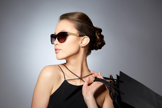 販売、ファッション、人、高級コンセプト - 灰色の背景にショッピング バッグと黒のサングラスで幸せな美しい若い女性