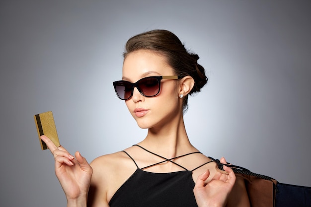 판매, 재정, 패션, 사람, 고급 개념 - 회색 배경 위에 신용카드와 쇼핑백을 넣은 검은색 선글라스를 쓴 행복한 아름다운 젊은 여성
