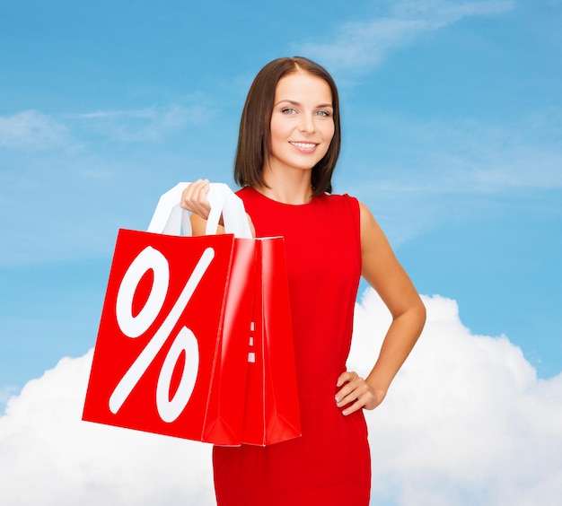 sale, gifts, christmas, holidays and people concept - smiling woman in red dress with shopping bags and percent sign over blue sky and white cloud background