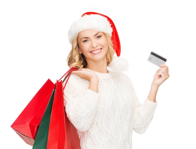 распродажа, подарки, рождество, рождественская концепция - улыбающаяся женщина в шляпе помощника Санты с сумками для покупок и кредитной картой