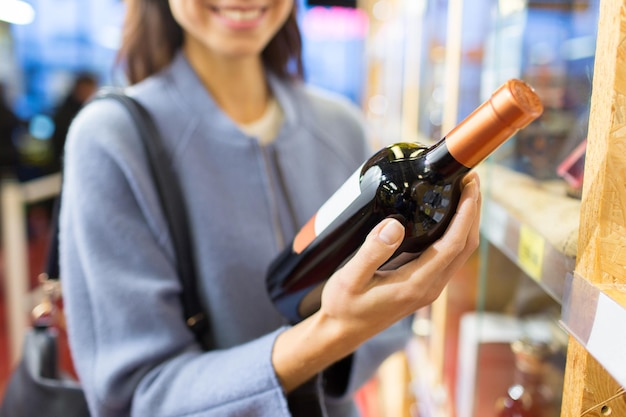 продажа, покупки, потребительство и концепция людей - счастливая молодая женщина выбирает и покупает вино на рынке или в винном магазине