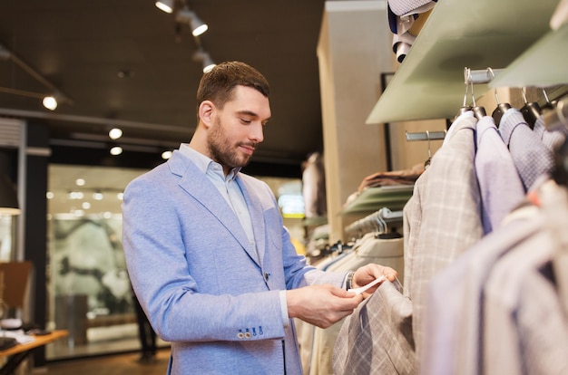 sale, shopping, fashion, style and people concept - elegant young man in jacket choosing clothes and looking at price tag in mall or clothing store