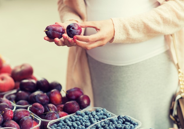 sale, shopping, pregnancy and people concept - close up of pregnant woman choosing plums at street food market