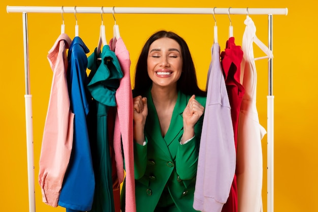 Sales and discounts concept Overjoyed young woman gesturing yes with joy standing among clothes in clothing rail