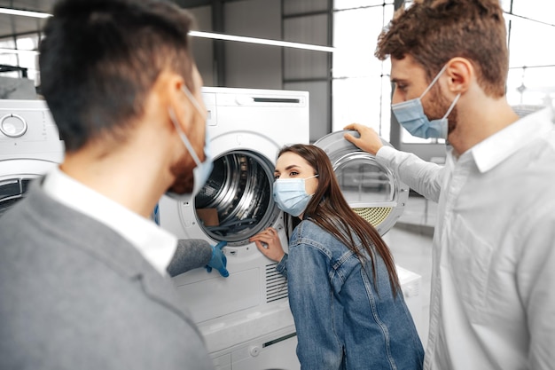 Salesman in hypermarket wearing medical mask demonstrates his\
clients a new washing machine