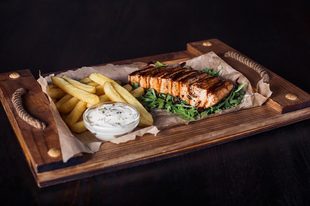 Salmon fillet steak with french fries on a wooden tray, beautiful serving, dark background.