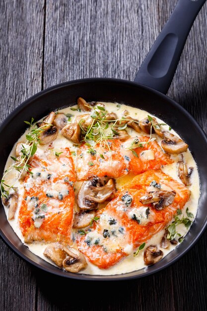 salmon fillet with creamy blue mold cheese sauce and mushrooms in pan on dark wooden table vertical view from above closeup