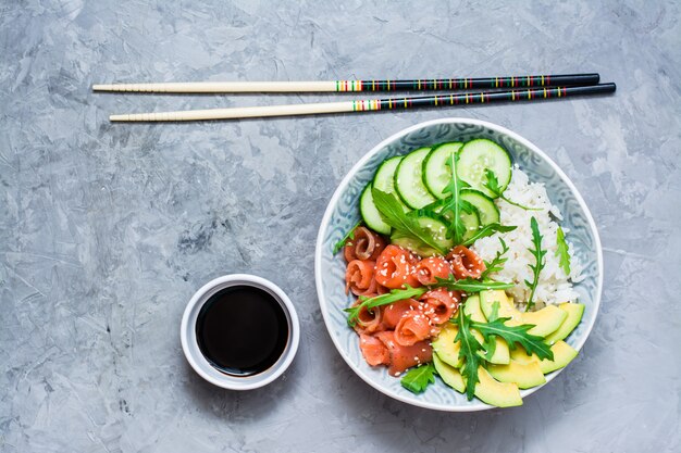 Photo salmon poke with avocado,  arugula and cucumber in a bowl, balsamic sauce and chopsticks.
