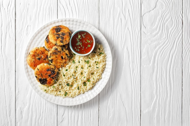 Salmon and spinach fish cakes with couscous and sweet chili sauce on a white plate on a wooden table, flat lay, free space
