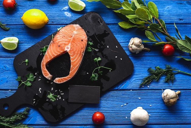 Salmon steak on a black cutting board and a blue wooden background, contrast, next to citrus vegetables and herbs