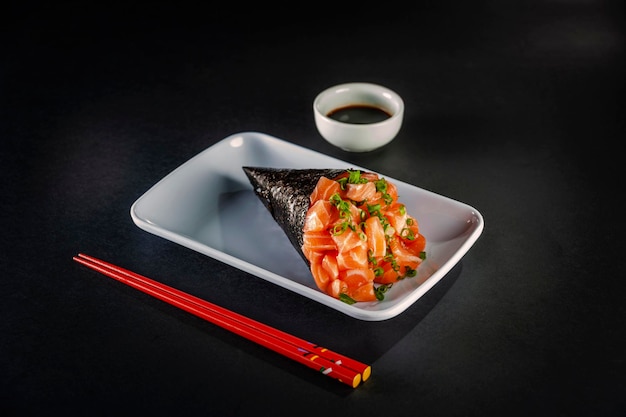 Темаки из лосося на белой тарелке на черном фоне с соевым соусом и палочками для еды.