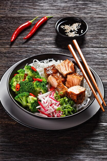 Salmon teriyaki with rice noodle broccoli radish and green onion in a bowl with chopsticks on a dark wooden table vertical view
