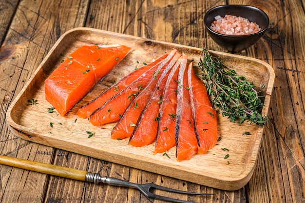 Salted salmon fillet slices in a wooden tray with thyme on wooden table. Top view.