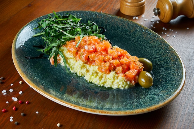 Salted salmon tartare on a scramble egg pillow with olives and
arugula on a blue plate on a wooden table. antipasti. delicious and
healthy breakfast food. close up