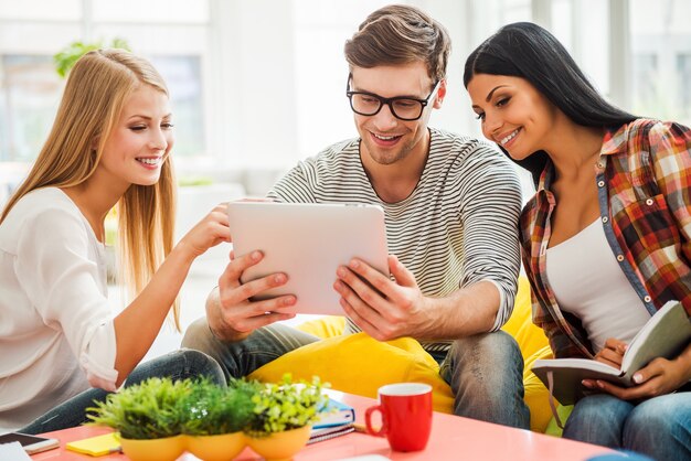 Samen aan een nieuw project werken. Drie gelukkige jonge mensen kijken naar digitale tablet en glimlachen terwijl ze op de kleurrijke zitzakken op kantoor zitten