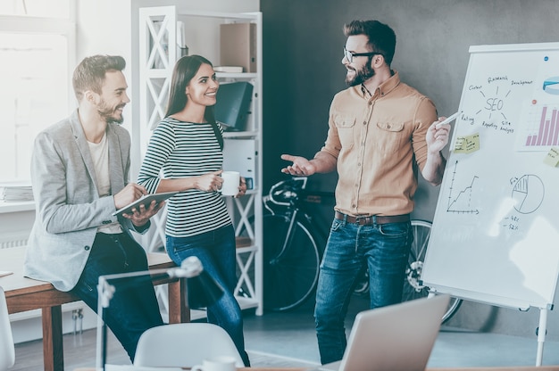 Samen een nieuw project bespreken. zelfverzekerde jonge man die in de buurt van het whiteboard staat en het met een glimlach wijst terwijl zijn collega's bij hem in de buurt staan