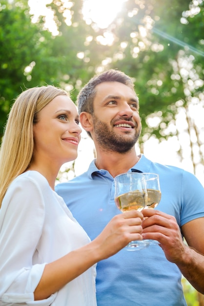 Samen genieten van tijd. Lage hoekmening van een gelukkig jong liefdevol stel dat een bril met witte wijn vasthoudt en wegkijkt terwijl ze buiten dicht bij elkaar staan