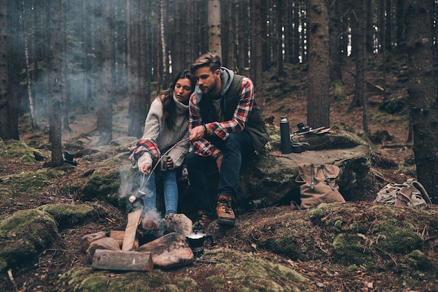 Samen genieten van tijd. Mooie jonge paar marshmallows roosteren boven een kampvuur zittend in het bos