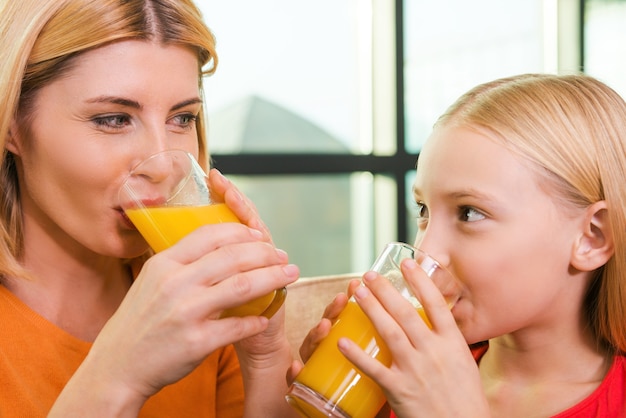 Samen genieten van vers sap. Vrolijke moeder en dochter drinken sinaasappelsap en kijken elkaar glimlachend aan terwijl ze samen op de bank zitten