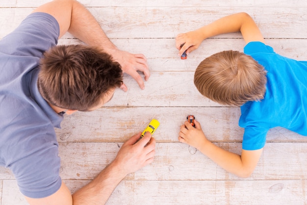 Samen spelen. Bovenaanzicht van vader en zoon die op de hardhouten vloer liggen en samen met speelgoedautootjes spelen
