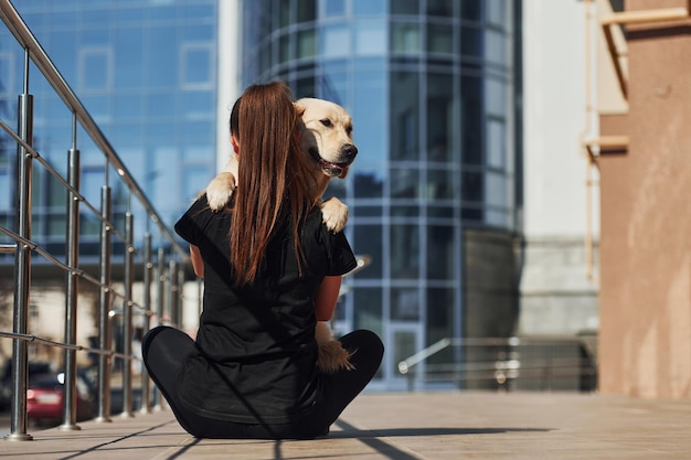 Samen zitten Jonge positieve vrouw heeft plezier met haar hond als ze buiten wandelt in de buurt van een bedrijfsgebouw