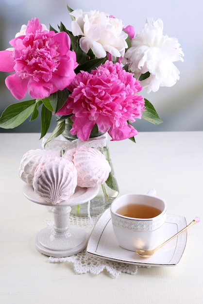 Samenstelling van mooie pioenrozen in vaas, thee in beker en marshmallow, op tafel, op lichte achtergrond