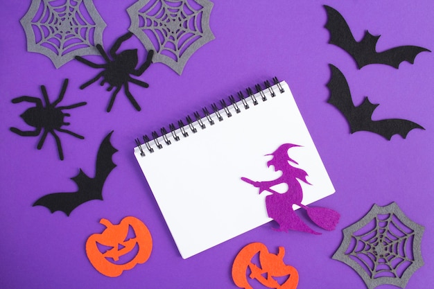 Samenstelling voor Halloween met wit notitieboekje, decoratieve heks, spinnen, pompoenen, spinnenwebben en vleermuizen op de paarse achtergrond. Ruimte kopiëren. Bovenaanzicht.