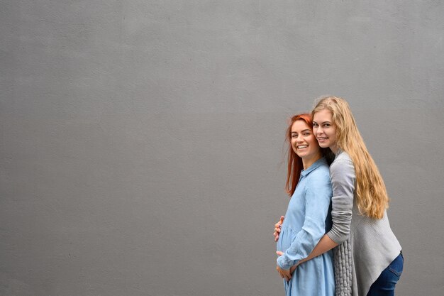 Samesex relationships A redhaired pregnant woman in a denim dress is standing against a gray wall her hand is resting under her tummy Her wife gently hugged the expectant mother from behind