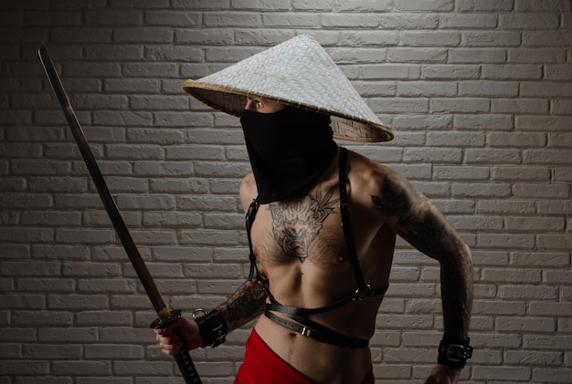 Samurai man in a red cape and an Asian triangular hat with tattoos and a katana sword in his hands against the background of a brick wall