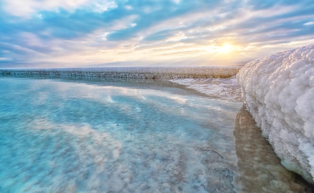 Sand completely covered with crystalline salt looks like ice or snow on shore of Dead Sea, turquoise blue water near, sky colored with morning sun distance - typical scenery at Ein Bokek beach, Israel