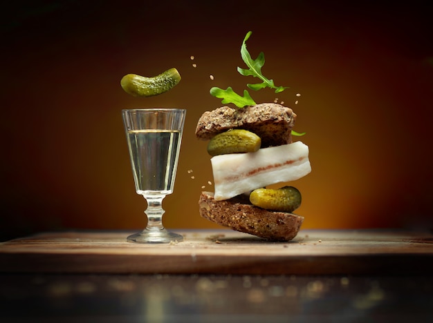 A sandwich in folk style with slice of bacon on brown background with flying ingredients pickles and arugula and a glass