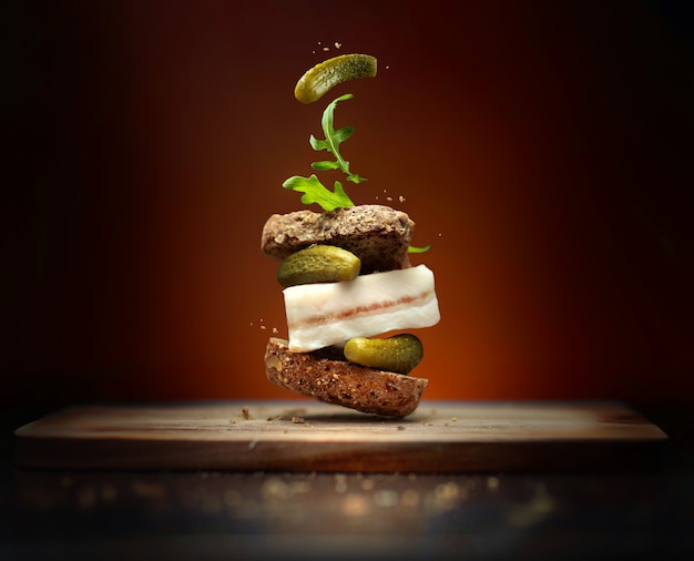 A sandwich in folk style with slice of bacon on brown background with flying ingredients pickles and arugula