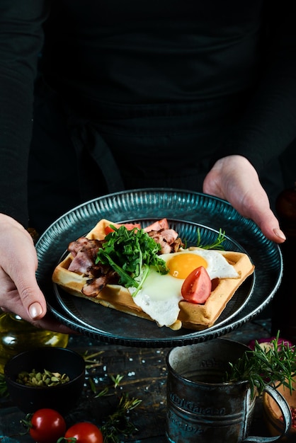 Sandwich met eitomaten en bacon op een bord in de handen van een chef-kok Op een zwarte achtergrond Belgische wafels