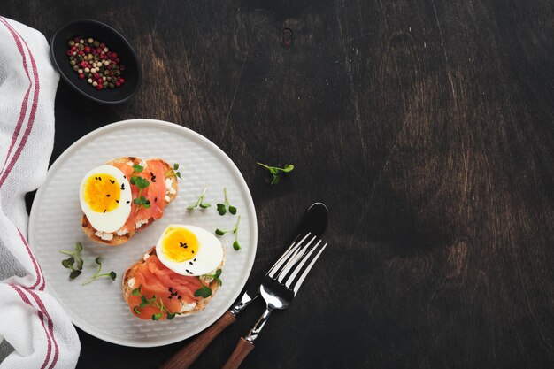 Sandwich met heerlijke toppings gerookte zalm eieren kruiden en microgreens radijs zwarte sesamzaadjes over witte plaat op oude houten tafel achtergrond Gezonde open sandwich superfood bovenaanzicht