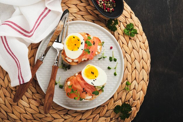 Sandwich met heerlijke toppings gerookte zalm eieren kruiden en microgreens radijs zwarte sesamzaadjes over witte plaat op oude houten tafel achtergrond Gezonde open sandwich superfood bovenaanzicht