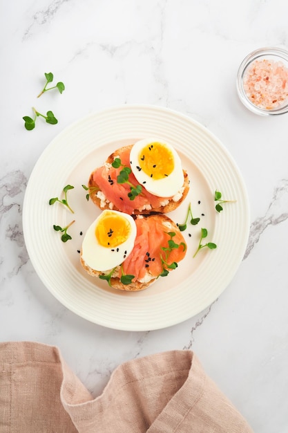 Sandwich met heerlijke toppings gerookte zalm eieren kruiden en microgreens radijs zwarte sesamzaadjes over witte plaat op witte marmeren tafel achtergrond Gezonde open sandwich superfood bovenaanzicht